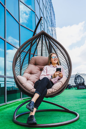 A fashionable woman relaxes in a cozy hanging chair on a spacious rooftop terrace, texting on her smartphone amid stunning modern architecture and clear blue skiesの写真素材