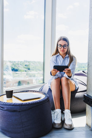 A young woman happily engages in her mobile gaming experience, surrounded by a stylish and contemporary living space that boasts beautiful scenic outdoor viewsの写真素材