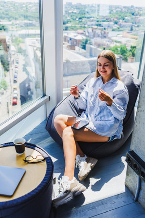 A young woman in a stylish outfit relaxes in a bean bag by the window, enjoying a snack and a beautiful cityscape viewの写真素材