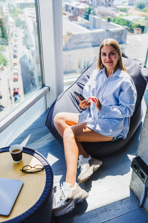 A young woman, dressed in a stylish blue striped shirt, finds a moment to relax in a cozy corner, savoring her coffee while using her smartphone in a bright and airy spaceの写真素材