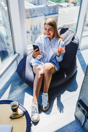 A cheerful young woman sits in a bean bag chair, enjoying her phone in a bright, sunny room overlooking the cityscape, radiating relaxation and modern lifestyle.の写真素材