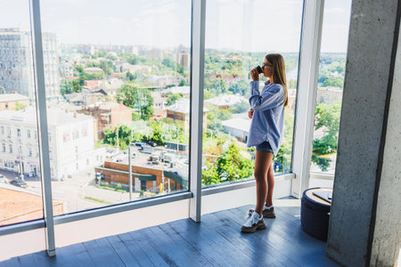 A young woman stands by large glass windows, capturing a vibrant urban cityscape outside with her camera, showcasing a moment filled with creativity and deep inspirationの写真素材