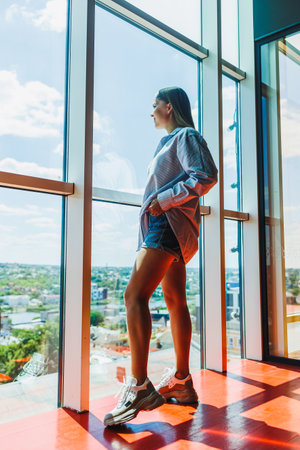 A stylish woman stands by a large window, lost in thought while gazing at the vibrant city skyline, dressed in casual but elegant attire that exudes charm and sophisticationの写真素材
