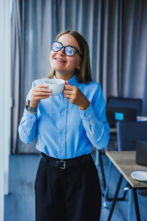 A cheerful young woman holds a steaming cup of coffee, stylish in a blue shirt and black trousers, radiating positivity and inspiring others in a vibrant officeの写真素材