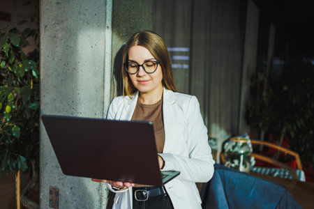 A young woman in stylish attire focused on her laptop in a contemporary workspace, surrounded by greenery and modern decor, embodying professionalism and creativity.の写真素材