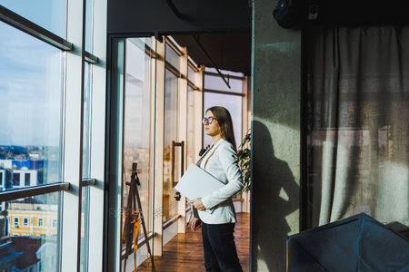 A stylish professional woman stands confidently by bright office windows, holding a sleek laptop that embodies her modern career ambitions in the business worldの写真素材
