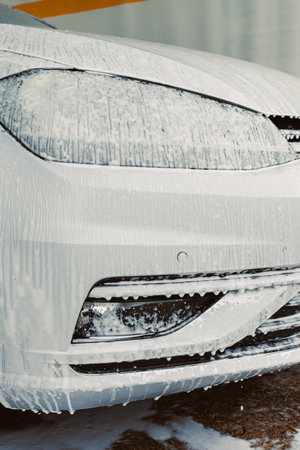 A pristine white car covered in foam at a car wash, showcasing the intricate details of the headlight and grille while being pampered with a thorough cleanの写真素材