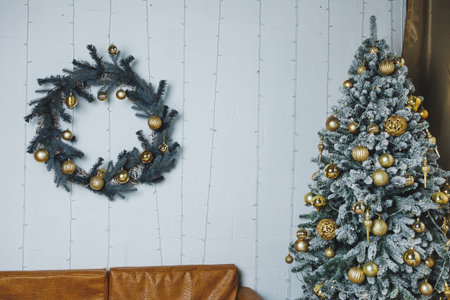 A festive living room scene showcasing a frosted Christmas tree adorned with golden ornaments and a stylish wreath on a white wall, perfect for holiday inspiration.の写真素材