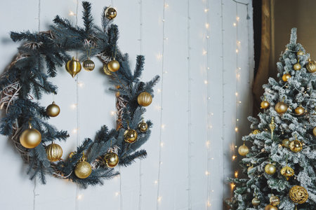 A beautifully decorated Christmas wreath with gold ornaments hangs on a white wall, accompanied by a snow-dusted tree adorned with golden baubles and twinkling lights.の写真素材