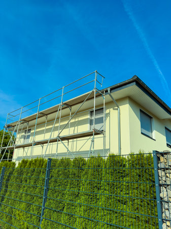 A contemporary house with scaffolding shows ongoing renovations, contrasting against a clear blue sky and lush greenery, embodying themes of construction and home improvementの写真素材