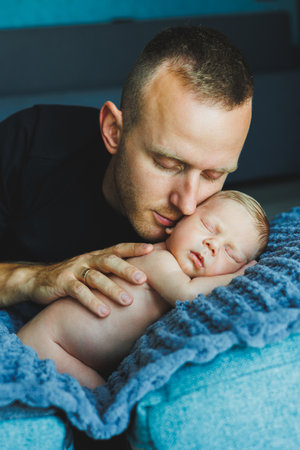 In a warm and joyful home, a loving father gently kisses his peacefully sleeping newborn, who is...