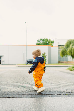 Smiling 2 year old boy in mudproof overalls and boots runs after the rain. Toddler in bright...