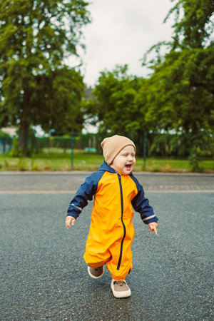 Cheerful boy 2 years old in yellow waterproof overalls walking along asphalt road on rainy day. Stylish boy in yellow waterproof jacket and rubber boots playing outdoors.の写真素材