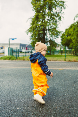 A cheerful child in a vibrant orange rain suit and cozy beige beanie joyfully walks along a wet...