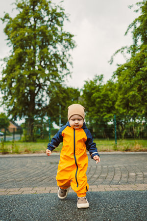 Cheerful boy 2 years old in yellow waterproof overalls walking along asphalt road on rainy day....