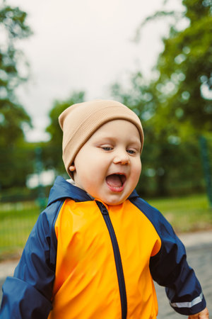 Cheerful baby enjoying outdoor playtime in a vibrant orange and blue raincoat and knit hat,...
