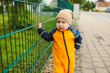 On a sunny day, a cheerful toddler in a bright orange rain suit and cozy beige hat joyfully explores a sturdy wooden fence, filled with laughter and playfulnessの写真素材