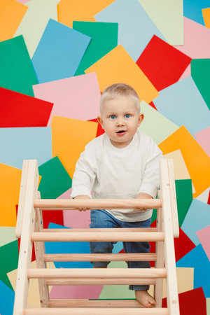 A joyful toddler in a white top and denim jeans climbs a sturdy wooden structure before a colorful wall, capturing the magic of childhood adventure and innocenceの写真素材