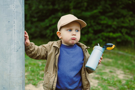 Adorable toddler wearing a cap, leaning against a post with a playful expression, holding a water...