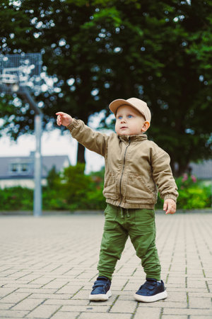 An energetic toddler in a stylish jacket and cute cap joyfully explores outdoors, pointing at...