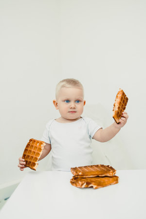 A curious toddler holds two waffle pieces with delight while several more are placed on the table,...