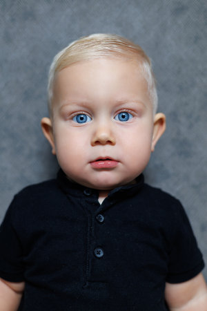 A cute young child with blue eyes and blonde hair is shown in a closeup, wearing a stylish black...