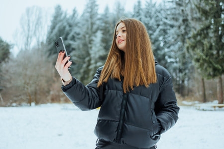A joyful young woman takes a fun selfie in a beautiful snowy landscape, wearing a stylish winter...