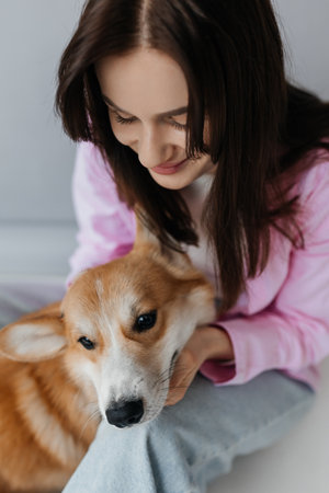 Happy brunette sitting next to corgi dog indoors. Cheerful girl in good mood stroking animal. Concept of friendship between man and pet.の写真素材
