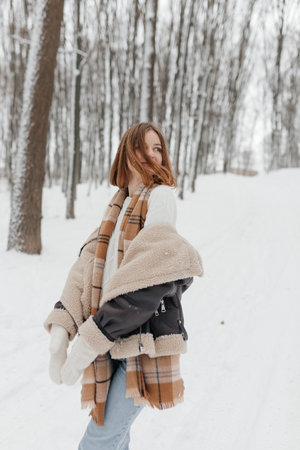 Happy smiling young woman wearing coat, scarf, and mittens enjoying winter weather while walking in...