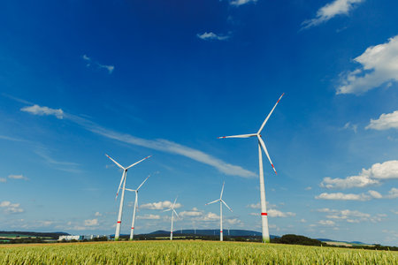 Wind turbines rotate against a backdrop of vibrant blue skies and fields, showcasing renewable...