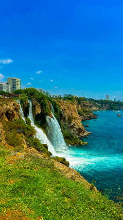 Stunning Duden Waterfalls plunging into the turquoise waters of the Mediterranean, surrounded by lush greenery and coastal city scenery, captured in bright daylight.の写真素材