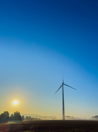 A tranquil landscape featuring a wind turbine at sunrise, bathed in golden light with a clear blue sky and soft mist, symbolizing renewable energy and nature's beauty.の写真素材