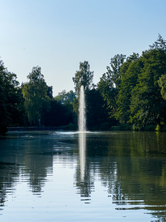 A peaceful park features a stunning fountain in a picturesque lake, surrounded by vibrant green trees under a clear blue sky, ideal for nature enthusiasts seeking tranquilityの写真素材