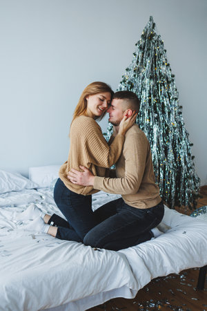 A loving couple enjoys an intimate moment on a bed, celebrating the holiday spirit with a beautifully decorated Christmas tree in the backgroundの写真素材