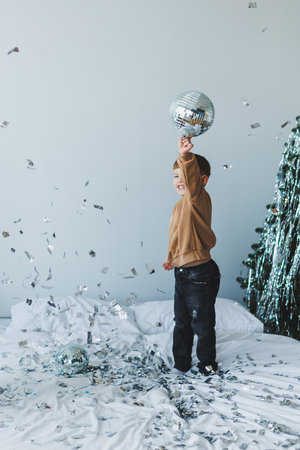 A cheerful child joyfully holds a shimmering disco ball among colorful confetti, near a decorated Christmas tree, capturing the essence of holiday celebration and joyの写真素材