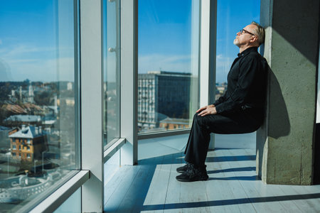 A man in elegant black attire sits by a window, enjoying the warm sunlight and a peaceful moment while gazing at a lively urban cityscape bustling with activityの写真素材