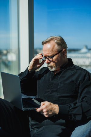 A focused man in stylish black attire is working on his laptop in a modern office, overlooking a...