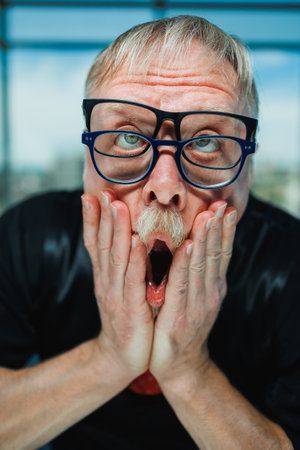 An older man with unique glasses features a surprised expression while holding his face, set against a vibrant backdrop. Perfect for conveying humor and surprise.の写真素材
