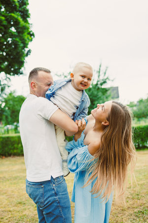 In a vibrant park, a joyful family captures a heartwarming scene as parents lift their smiling baby high, surrounded by greenery under a sunny sky, creating memoriesの写真素材