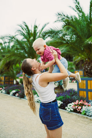 In a sunny garden with palm trees and colorful flowers, a joyful mother lifts her baby high, creating a heartwarming scene of love, happiness, and togethernessの写真素材