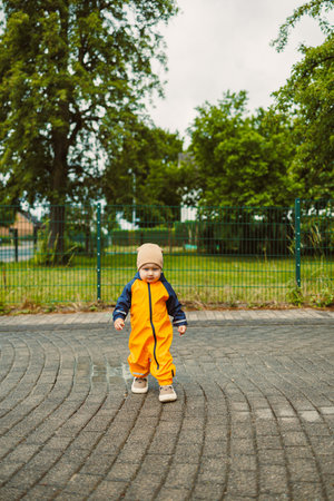 A cheerful toddler in an orange rain suit and beige hat is joyfully exploring a park on a cloudy day, with lush grass and tall trees creating a perfect backdrop for funの写真素材