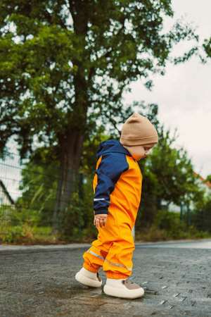 On a rainy day, a young child in a bright orange rain suit and colorful beanie joyfully leaps into puddles, embodying the essence of playful childhood explorationの写真素材