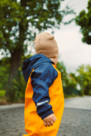 A young child in colorful rain gear walks along a scenic path, surrounded by lush greenery, joyfully exploring nature on a cool, overcast day, embodying innocent childhoodの写真素材