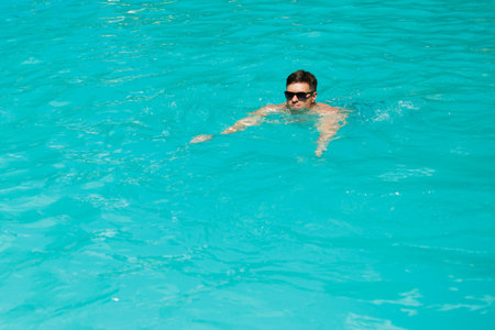 A joyful young man happily swims in a stunning turquoise pool, soaking in the warm sun while savoring the refreshing water on a delightful, perfect sunny day that feels just rightの写真素材