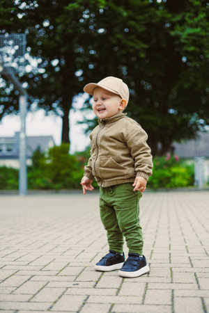 A cheerful toddler dressed in a cozy sweater and cap stands on a paved surface, smiling happily amidst green trees, embodying the spirit of childhood play and joy.の写真素材