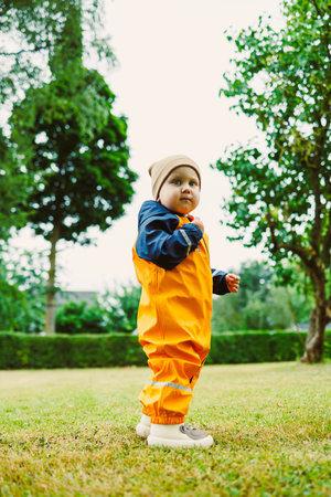 A cheerful toddler in a vibrant orange rain suit and matching hat enjoys a sunny day outdoors,...