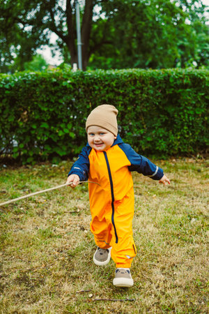 A joyful toddler in a bright orange rain suit and beige hat runs through green grass, capturing...