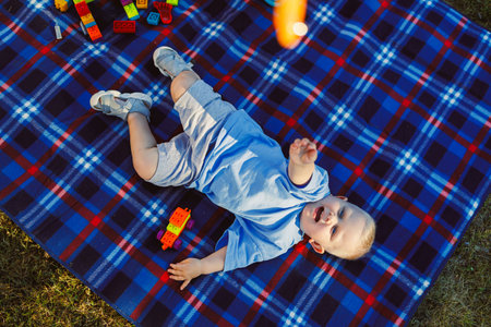A cheerful toddler lies on a bright blanket, happily playing with colorful blocks outdoors. This...