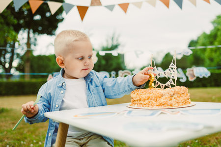 A cheerful young child interacts with a beautifully decorated cake at an outdoor birthday party,...