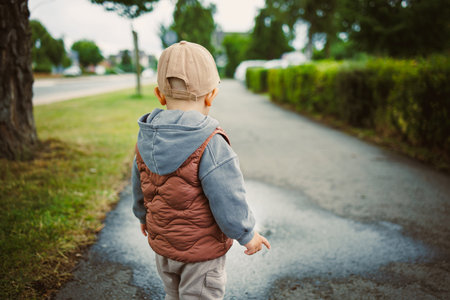 A young child in a cozy sweater and stylish vest strolls along a picturesque, treelined pathway,...